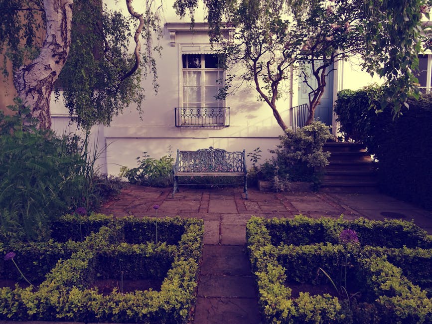 A quaint backyard garden featuring a paved stone patio area with a decorative metal garden bench positioned against a white exterior wall of a house. The garden is bordered by well-maintained, neatly 