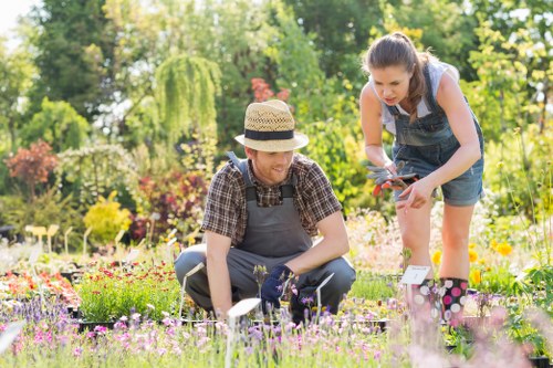 In a well-maintained garden in Blackheath, two individuals are engaged in gardening activities amidst a lush outdoor setting. The garden features a vibrant array of flowering plants with various colours, including pinks, yellows, and reds, planted in neatly edged flower beds. The foreground showcases a lush, green lawn with dense grass, appearing healthy and well-kept. In the background, there are tall trees and a leafy hedge, providing a natural backdrop and privacy to the outdoor space. One person is kneeling on the grass, wearing a straw hat, a checkered shirt, and gardening gloves, actively tending to the plants with a hand tool. The other stands next to them, dressed in a sleeveless top and denim shorts, leaning forward slightly while holding gardening shears, assisting or inspecting the work. The garden receives ample sunlight, indicating a bright, clear day, and the overall scene depicts a tidy, cultivated outdoor environment suitable for various gardening services offered by Garden Maintenance Blackheath, contributing to the area's charming, natural landscape and outdoor leisure space.